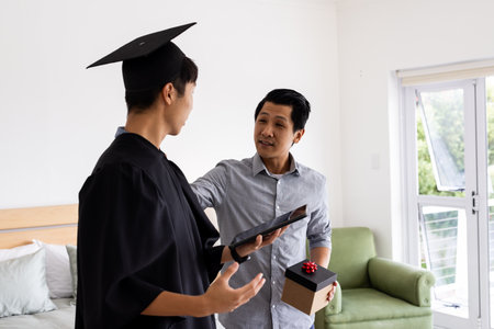 At home, father giving gift to son in graduation gown, celebrating achievement. Family, celebration,...