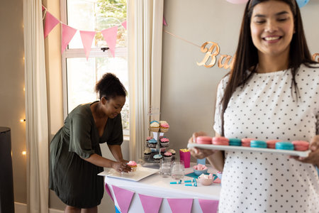 Women preparing colorful macarons and cupcakes for festive baby shower celebration. Baking, dessert,...