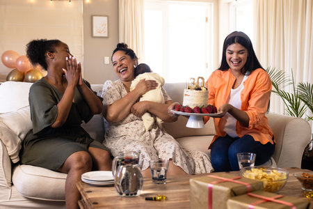 Celebrating birthday, women laughing with cake and baby on cozy living room sofa. Celebration,...