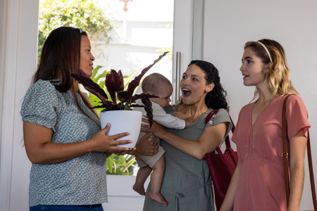 Women greeting each other at home, holding baby and houseplant, smiling warmly. Motherhood,...