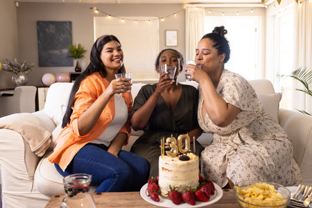 Celebrating at home, diverse women enjoying drinks with birthday cake and snacks. Celebration, party, friendship, togetherness, happiness, leisureの写真素材