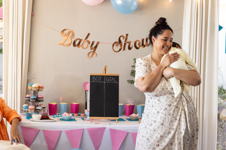 Smiling woman holding baby at baby shower celebration with decorations and cupcakes. Motherhood,...