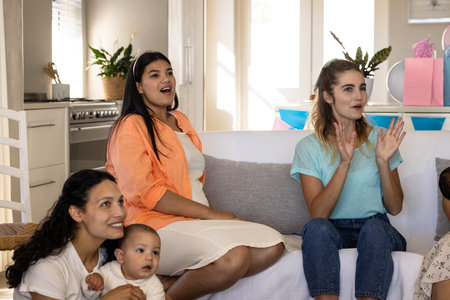 Women and baby sitting on couch, joyfully watching something exciting at home. Family, motherhood, bonding, parenting, entertainment, relaxationの写真素材