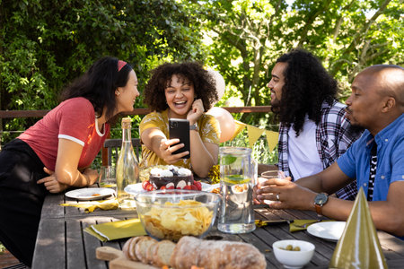 Celebrating outdoors, friends laughing and taking selfie at festive table with snacks. party, celebration, gathering, joyful, technology, socializingの写真素材