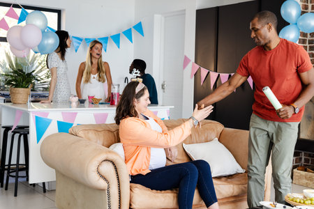African American man handing gift to pregnant woman at home baby shower celebration. happiness, love, family, togetherness, community, pregnancyの写真素材
