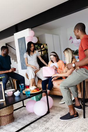 Friends celebrating at home, young woman opening gift with excitement and joy. Celebration,...