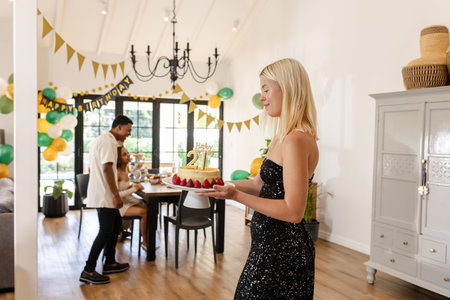 Blonde woman carrying birthday cake while friends celebrate at home party. Celebration, enjoyment, togetherness, festive, gatheringの写真素材