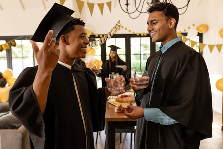 Graduation celebration, two young men in caps and gowns toasting with drinks. achievement, success, ceremony, educationの写真素材