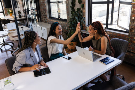 Colleagues high-fiving at office table, celebrating success with laptops and tablets. Teamwork, collaboration, business, technology, meeting, communicationの写真素材