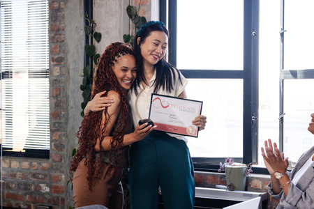 Asian woman holding certificate hugging colleague in office, celebrating achievement. Congratulations, teamwork, success, recognition, diversity, awardsの写真素材
