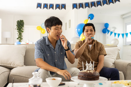 Celebrating birthday, two men enjoying cake and blowing party horns at home. Celebration, happiness, friendship, fun, dessert, indoorの写真素材