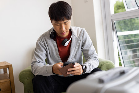 Young man using smartphone at home, wearing headphones, sitting on green chair. Technology, communication, listening, relaxation, leisure, modernの写真素材