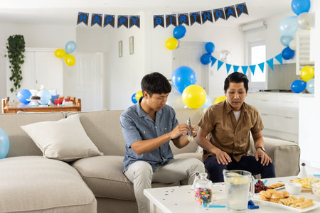Celebrating birthday, two men sitting on couch with decorations and snacks. Celebration, party, friendship, happiness, fun, enjoymentの写真素材