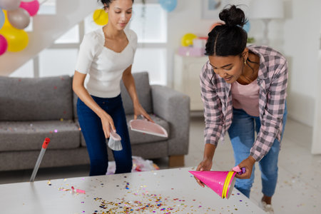 Two women cleaning up party decorations and confetti in living room. Cleanup, party aftermath, tidying, mess, domestic, organizationの写真素材