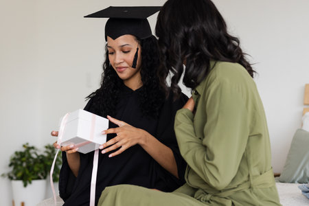 African American graduate receiving gift from proud mother during celebration at home. Graduation, achievement, family, college, success, happinessの写真素材
