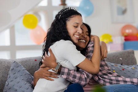 Two happy girls hugging on couch at home during birthday celebration. Friendship, happiness, bonding, indoors, festive, joyの写真素材