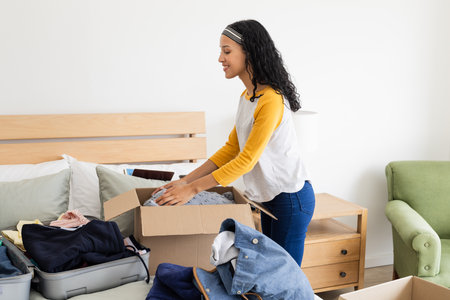 Woman packing clothes into cardboard box on bed, preparing for moving day. Relocation, organizing,...