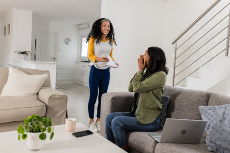 African American woman surprising friend with letter while sitting on sofa at home. Friendship, happiness, emotions, surprise, celebration, leisureの写真素材