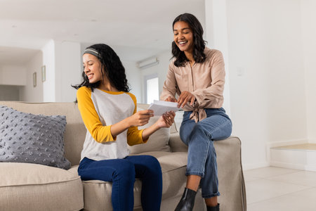Mother and daughter sitting on sofa, reading letter together, smiling happily. Family, bonding, relationship, parenting, communication, emotionsの写真素材