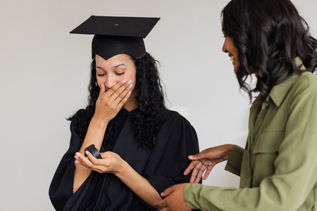 Graduate in cap and gown surprised with gift, smiling woman congratulating her. Graduation, achievement, celebration, commencement, education, successの写真素材