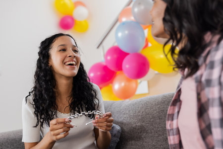 Chatting with friend on couch, woman holding bracelet, colorful balloons behind. Friendship, celebration, leisure, relaxation, jewelry, partyの写真素材