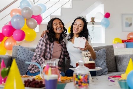 Exchanging gifts and smiling, two African American women celebrating birthday on couch. Celebration, happiness, friendship, presents, joy, homeの写真素材