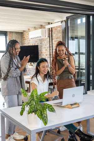 Team celebrating success at office, three women clapping and smiling at laptop. Teamwork, celebration, achievement, business, collaboration, workplaceの写真素材