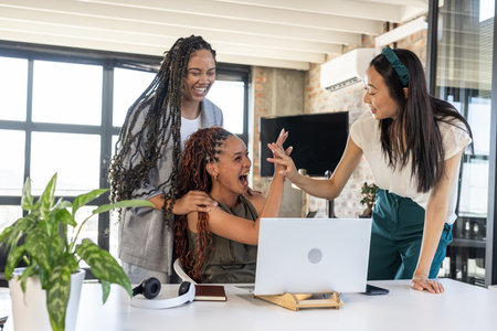 Celebrating success, diverse businesswomen high-fiving at office desk with laptop. Teamwork, collaboration, empowerment, leadership, diversity, corporateの写真素材