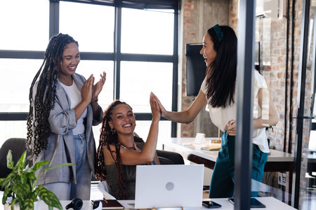 Celebrating success, diverse businesswomen high-fiving in modern office with laptop. teamwork, achievement, diversity, collaboration, motivation, celebrationの写真素材