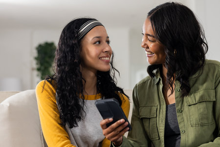 Smiling, African American mother and daughter looking at smartphone together at home. Family,...