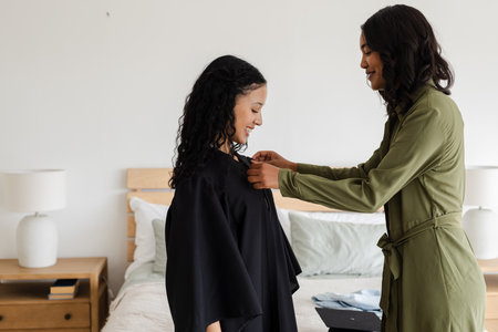 Mother helping daughter adjust graduation gown in cozy bedroom setting. family, celebration, education, milestone, achievement, parentの写真素材