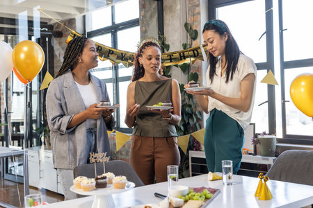 African American and Asian women enjoying birthday celebration at office with cake. diversity, corporate, team, workplace, friendship, eventの写真素材