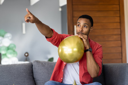 Man blowing gold balloon on couch, pointing excitedly at something indoors. Celebration, party, enthusiasm, decoration, happiness, funの写真素材