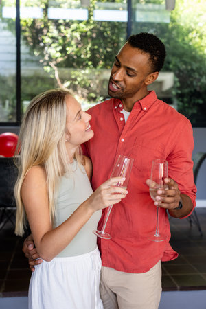 Couple celebrating at home, holding champagne glasses and smiling at each other. Celebration, romance, relationship, happiness, toast, loveの写真素材
