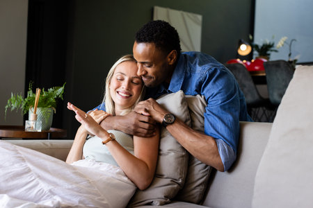 African American man hugging smiling woman on couch at home, enjoying togetherness. couple, relationship, bonding, love, lifestyle, living roomの写真素材