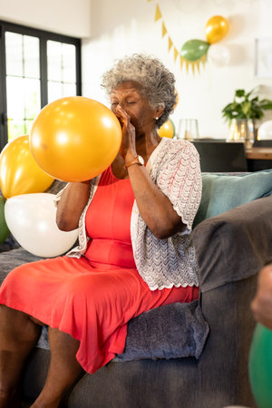 Elderly woman inflating balloon on couch, preparing for festive celebration. Party, senior, decorating, event, jubilantの写真素材