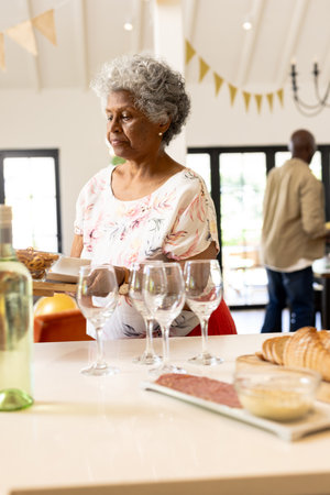 Elderly woman preparing table with wine glasses and snacks for celebration at home. hospitality, gathering, festivities, dining, senior, preparationの写真素材