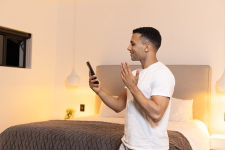 Man in bedroom video chatting on smartphone, smiling and gesturing happily. Communication, technology, connection, conversation, virtual, homeの写真素材