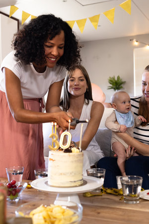 Smiling woman cutting birthday cake with friends and baby at home celebration. party, friendship, family, gathering, joyful, happinessの写真素材