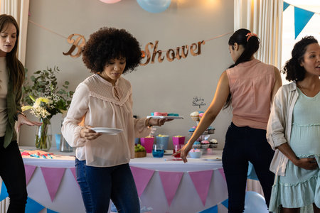 Women enjoying baby shower, holding plates and chatting near decorated table indoors. Celebration, maternity, gathering, friendship, party, balloonsの写真素材