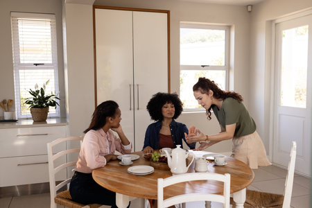 Friends enjoying tea and conversation at home around wooden kitchen table. Friendship, gathering, socializing, beverage, chatting, lifestyleの写真素材