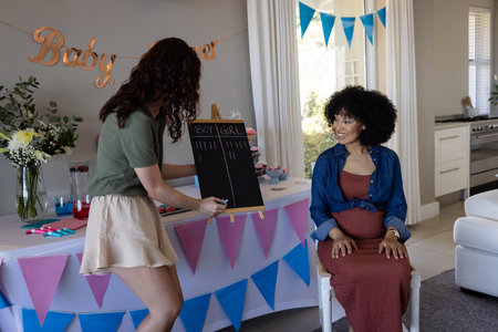 Woman writing on chalkboard at baby shower while pregnant friend smiles. Celebration, maternity, friendship, fun, expecting, joyの写真素材