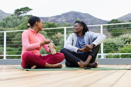 African American friends twisting open reusable water bottle sitting on yoga mats on wooden terrace....