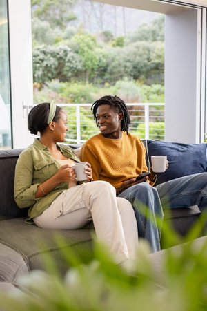 African American couple chatting on dark gray sofa in living room holding ceramic mugs and tablet. Coziness, intimacy, relaxation, hospitality, home decor, lifestyle, modernの写真素材