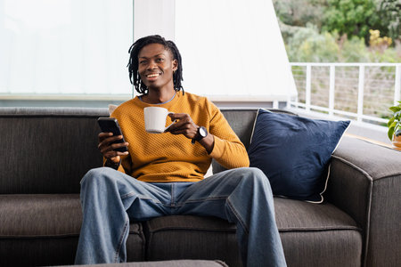 Mid adult African American man sitting on sofa holding mug and checking smartphone at home. Casual,...