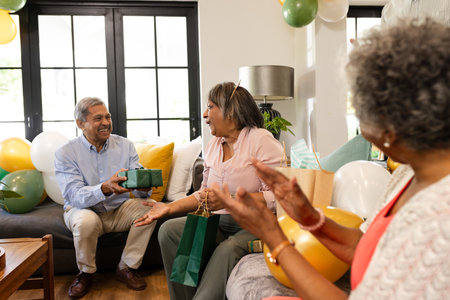Elderly man giving gift to woman at home, celebrating with friends and balloons. Celebration, party,...