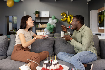 Friends laughing and sharing birthday cake together on living room sofa. Celebration, friendship, gathering, dessert, happiness, partyの写真素材