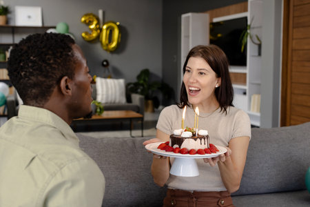 Woman holding birthday cake with candles, smiling at man in living room. Celebration, birthday party, happiness, togetherness, festive, joyfulの写真素材