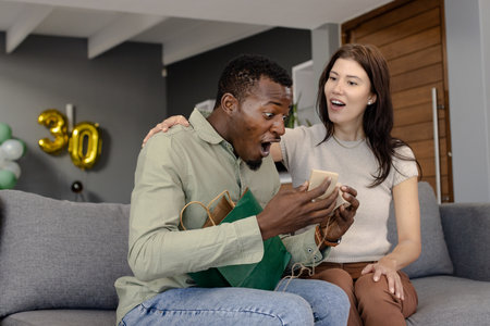 Celebrating birthday, surprised man opening gift with friend on living room sofa. Celebration,...