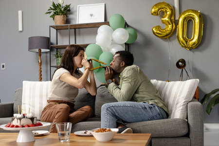 Couple celebrating birthday at home, blowing party horns on couch, feeling joyful. Celebration, happiness, festive, balloonsの写真素材
